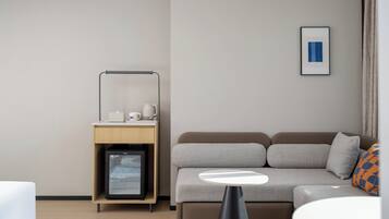 A modern hotel room with a sofa, a small table, a minibar, and a framed picture on the wall.