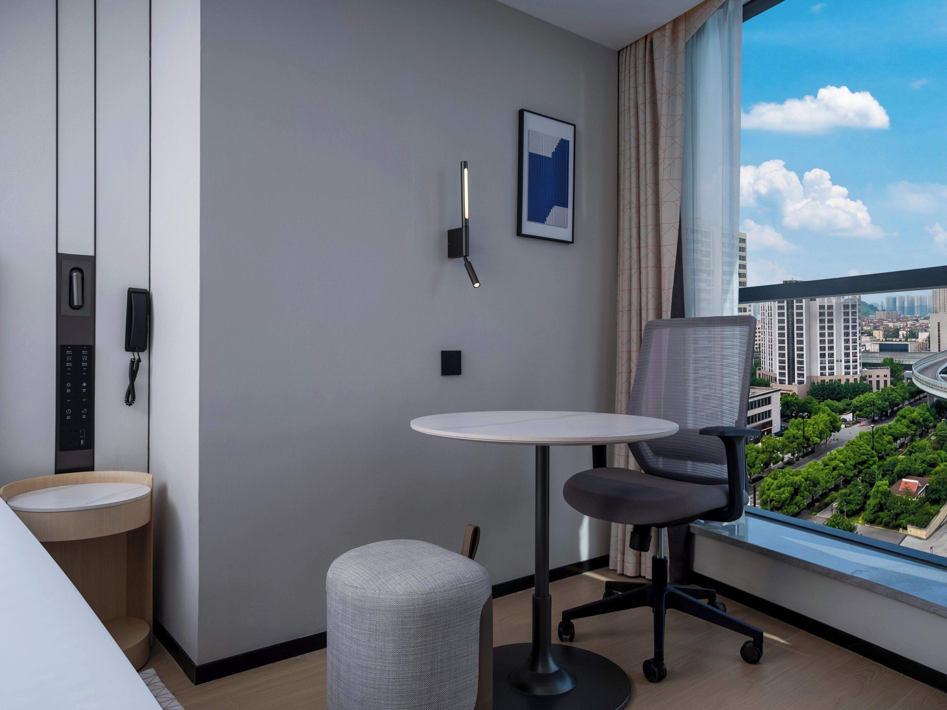 A modern hotel room with a round table, a cushioned chair, a phone, a picture on the wall, and a window with a city view.