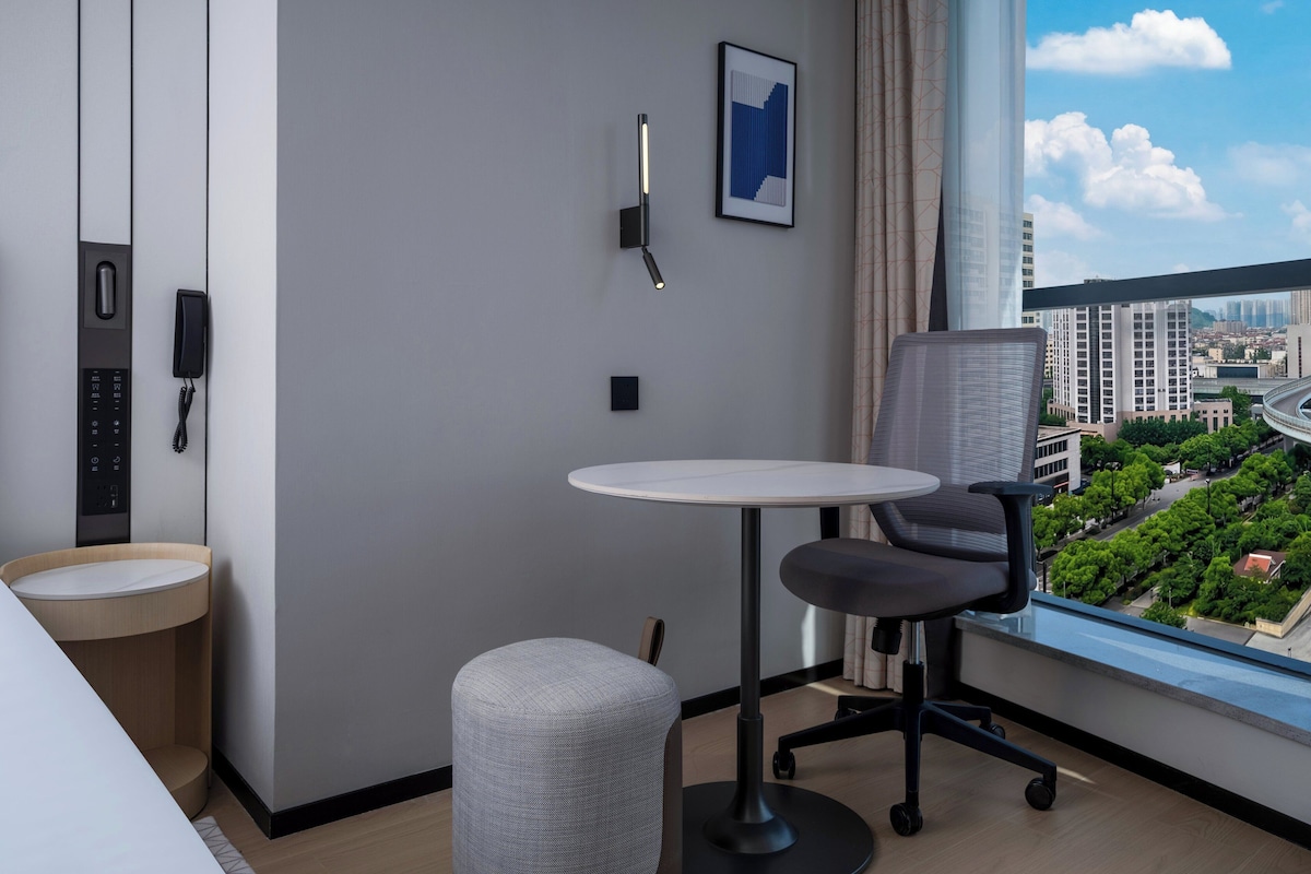 Habitación de hotel moderna con una mesa redonda, una silla acolchada, un teléfono, un cuadro en la pared y una ventana con vista a la ciudad.
