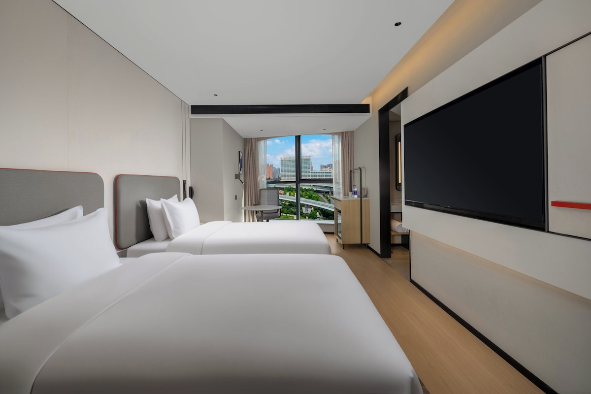 Habitación de hotel con dos camas, televisión, escritorio y vista a la ciudad.