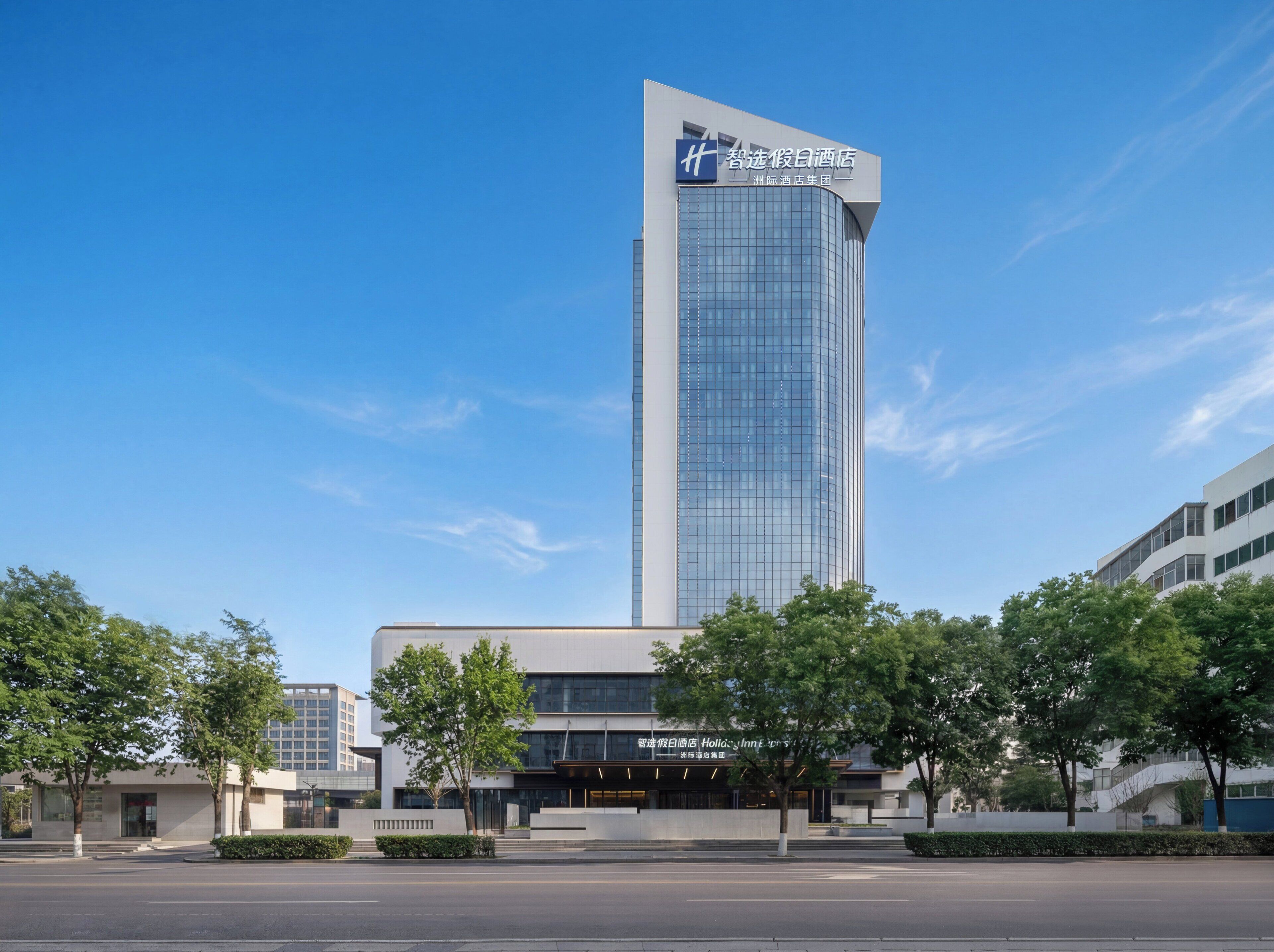 A modern hotel building with a prominent glass facade, a sign indicating it is part of the Zhujiajie International Hotel Group, and a well-maintained surrounding area with trees and a clear sky.