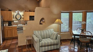Un salón acogedor con un sofá a rayas, una mesa de comedor con sillas, una zona de cocina con armarios de madera y un ventanal con persianas.