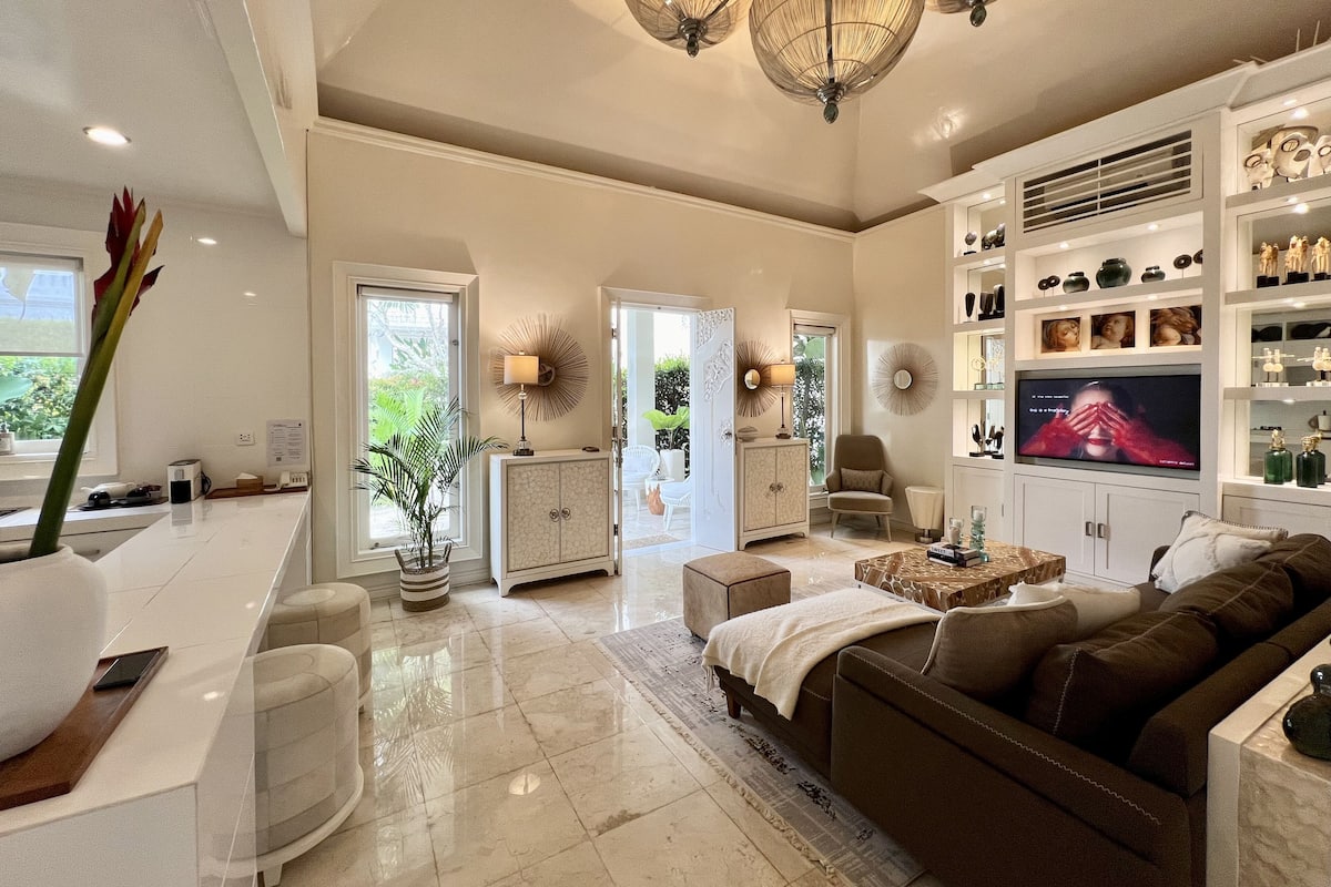 A spacious living room with a large brown sofa, a coffee table, and a television. There is a cabinet with decorative items and a plant in the corner.