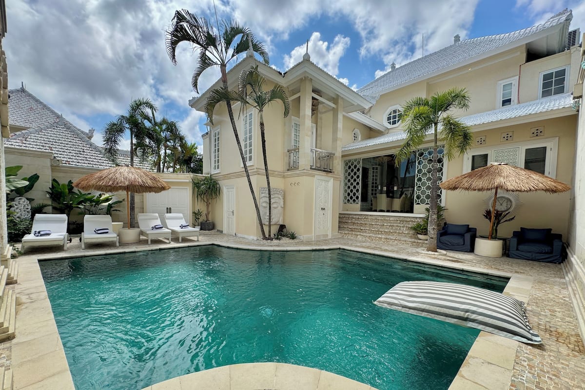 A swimming pool with lounge chairs and umbrellas, surrounded by palm trees and a building with a tiled roof.