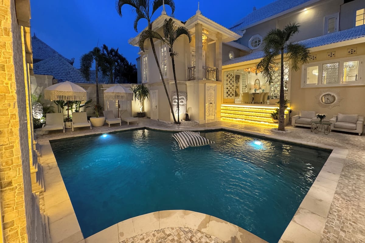 A swimming pool with a stone deck, palm trees, and a well-lit outdoor seating area.