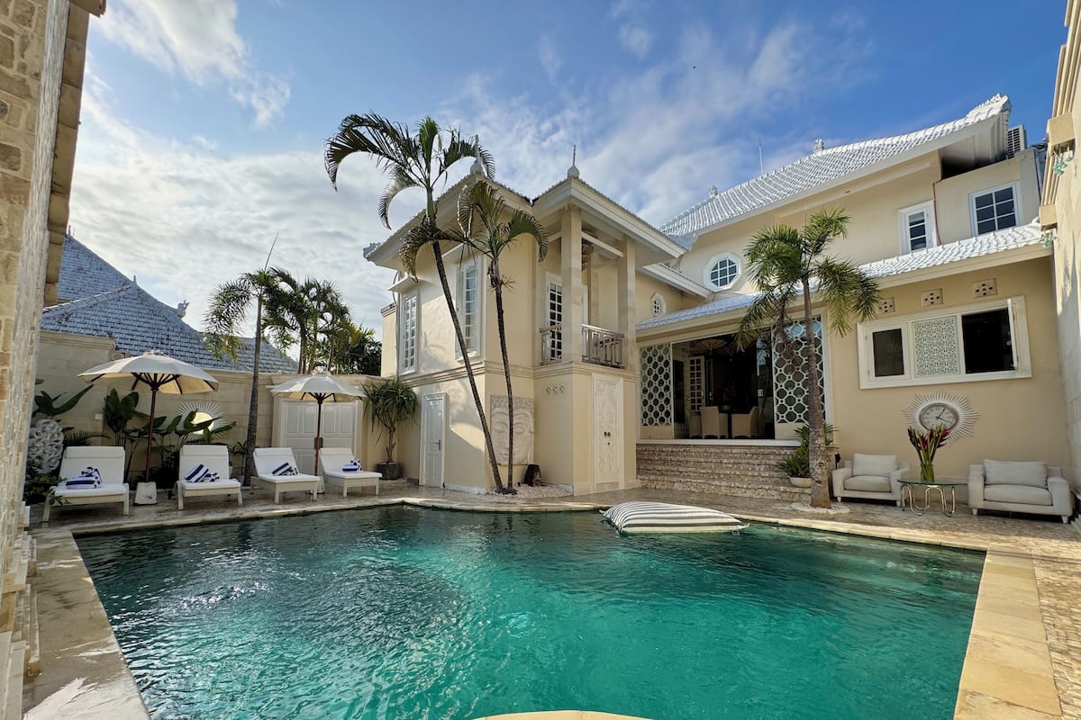 A pool area with lounge chairs and umbrellas, a large house with a tiled roof, and palm trees.