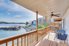 A wooden deck with a ceiling fan, overlooking a calm lake with docks and buildings in the background. - Waterfront Condo w/ Deck on Lake of the Ozarks! (Lake Ozark)
