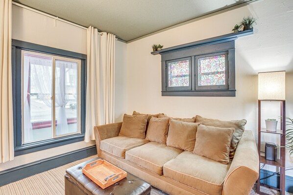 A living room with a beige sofa, a coffee table, a window with curtains, and a stained glass window above the sofa. - Charming Minneapolis Home w/ Deck, 5 Mi to Central (Minneapolis)