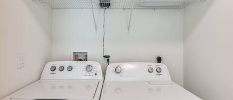 A laundry room with two Whirlpool washers and dryers, a white laundry basket on a shelf, and a power outlet on the wall.