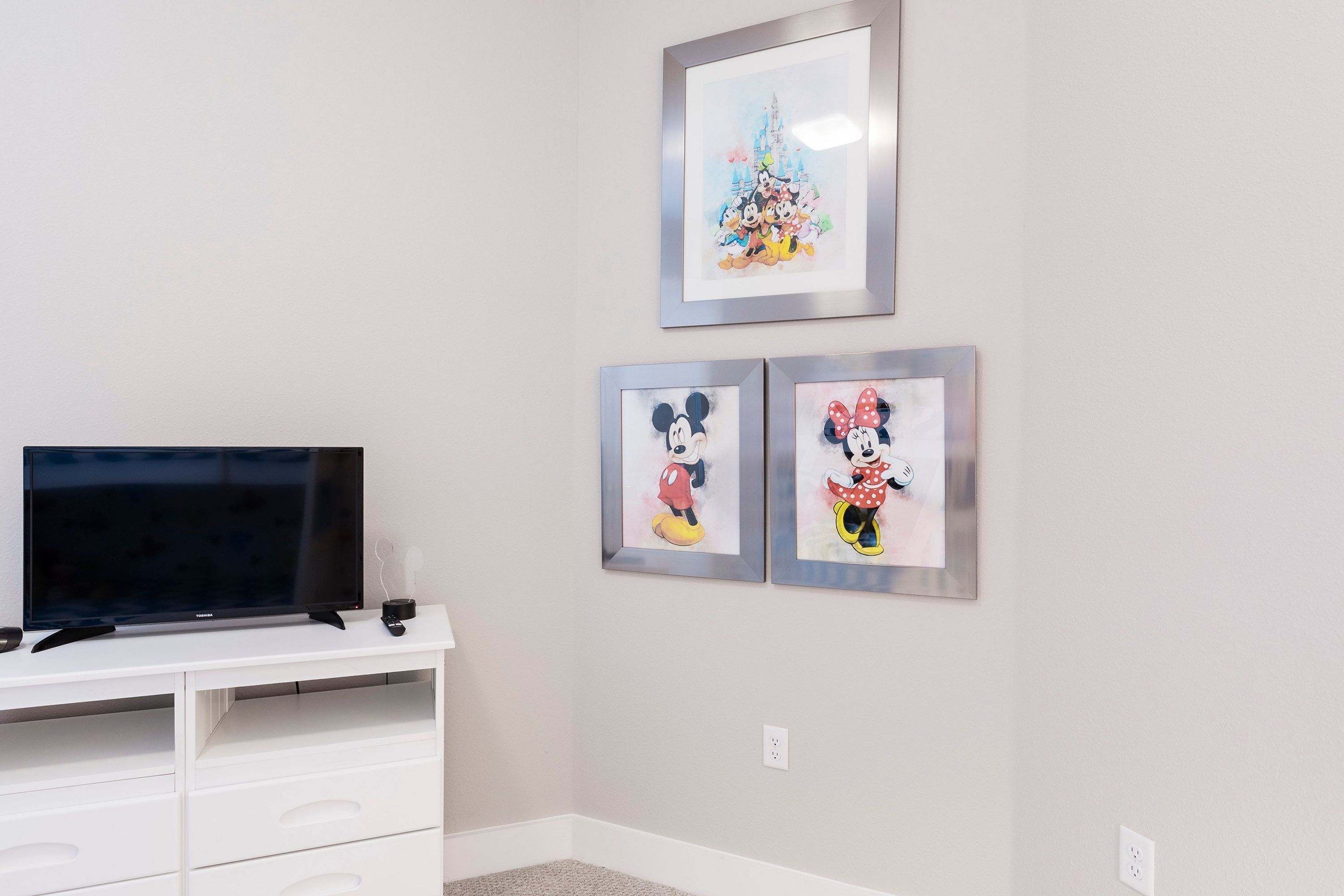 Ett rum med en platt-TV placerad på en vit TV-bänk, två inramade tryck med Disneyfigurer på väggen och ett rum med heltäckningsmatta.