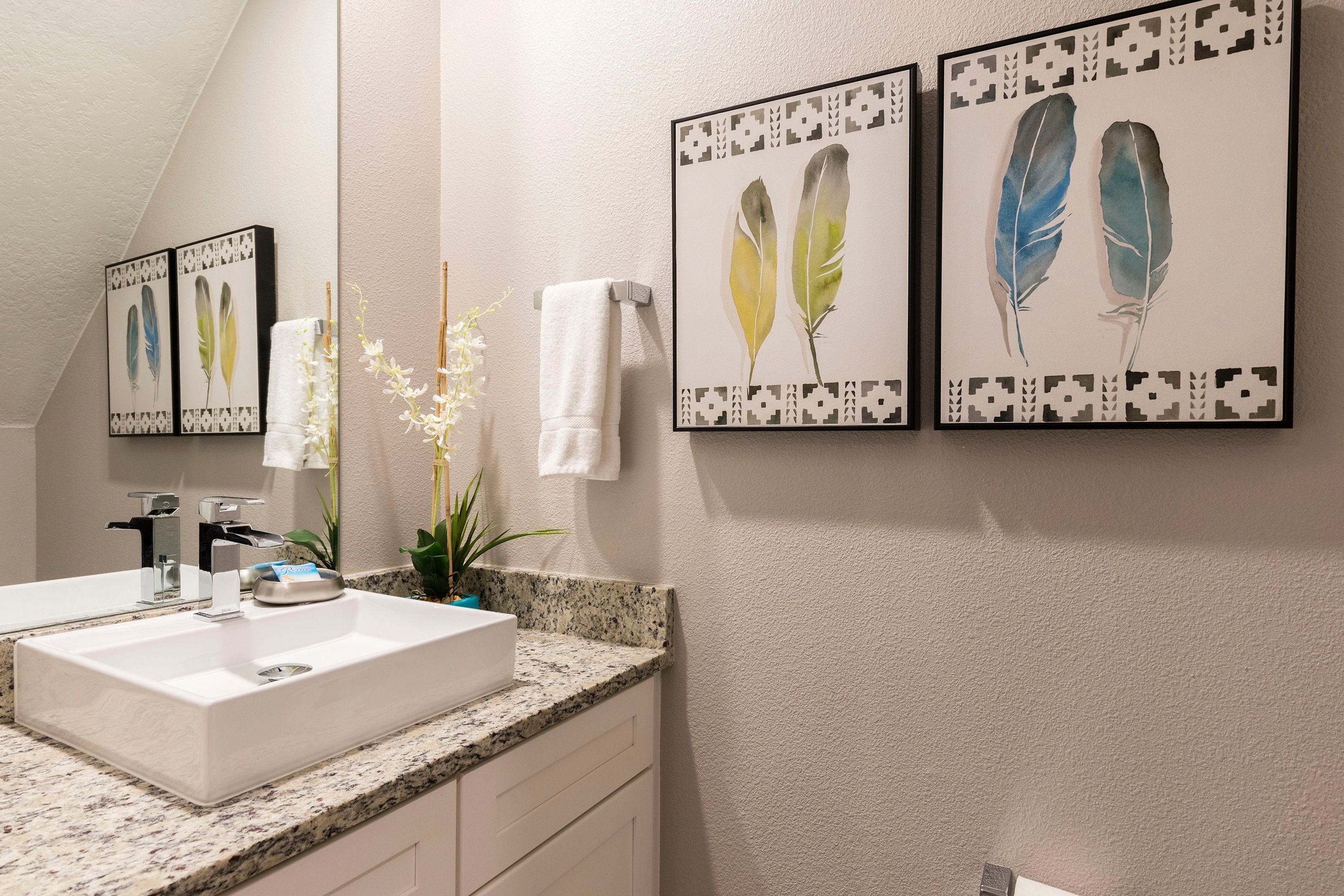 Une salle de bain avec un lavabo blanc, un miroir et une œuvre d’art représentant des plumes, encadrée et accrochée au mur.