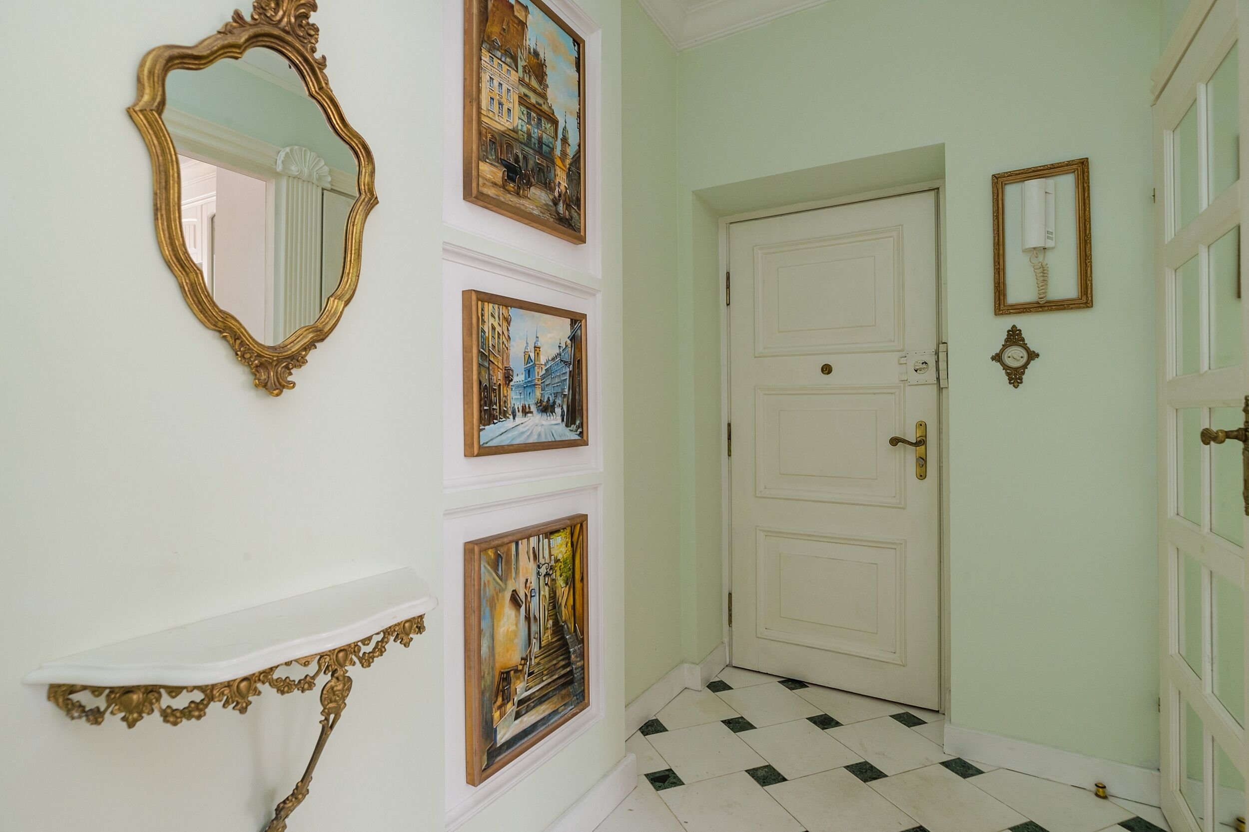A hallway with a white door, a gold-framed mirror, and three framed paintings on the wall.