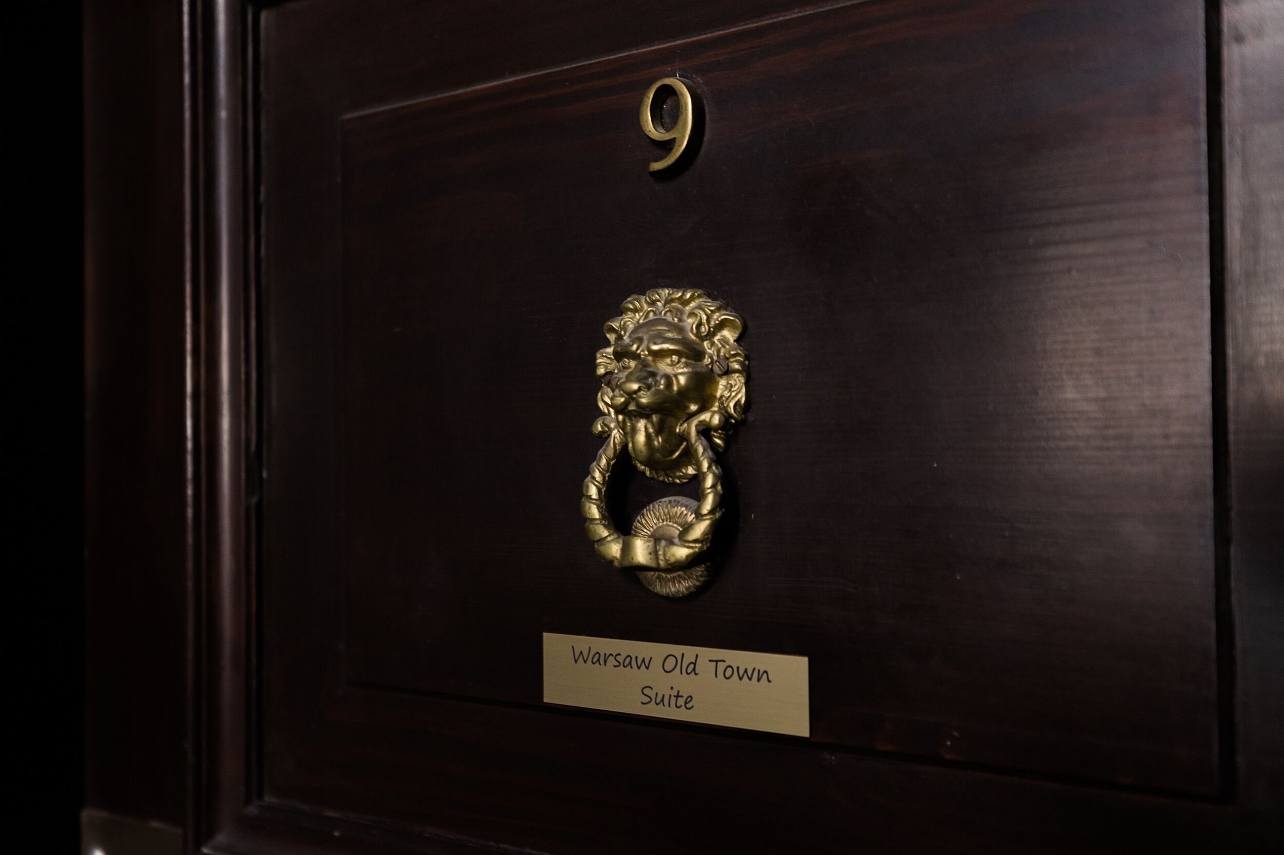 Uma porta de madeira escura com um aldabinha em forma de cabeça de leão em latão e uma placa com a inscrição 'Warsaw Old Town Suite'.
