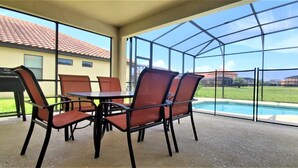 A covered patio with a table and chairs, a grill, and a view of a swimming pool and houses. - Grand Superior 4BD Pool Home at Disney Universal (Davenport)
