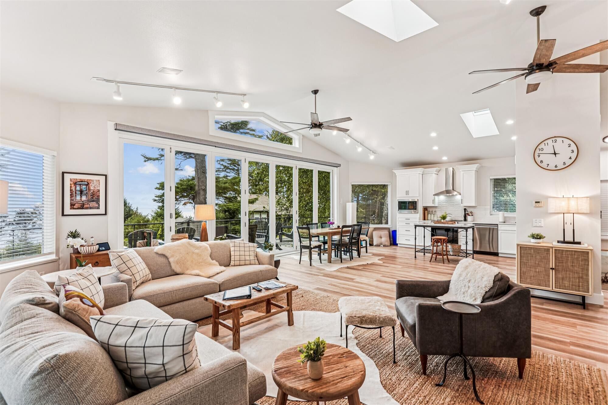 A spacious living room with a large sofa, coffee table, and a dining area with chairs. There is a kitchen with white cabinets and a ceiling fan.