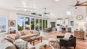 A spacious living room with a large sofa, coffee table, and a dining area with chairs. There is a kitchen with white cabinets and a ceiling fan.