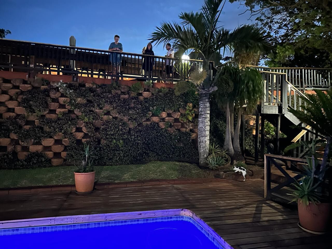 Een zwembadgedeelte met een houten terras, een stenen muur met beplanting en een trap naar een balkon.