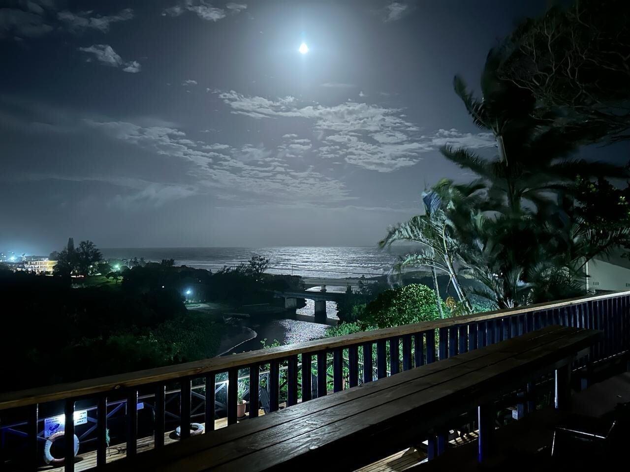 Vista nocturna desde un balcón con vista a una playa, un puente y palmeras.
