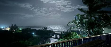 Vista nocturna desde un balcón con vista a una playa, un puente y palmeras.