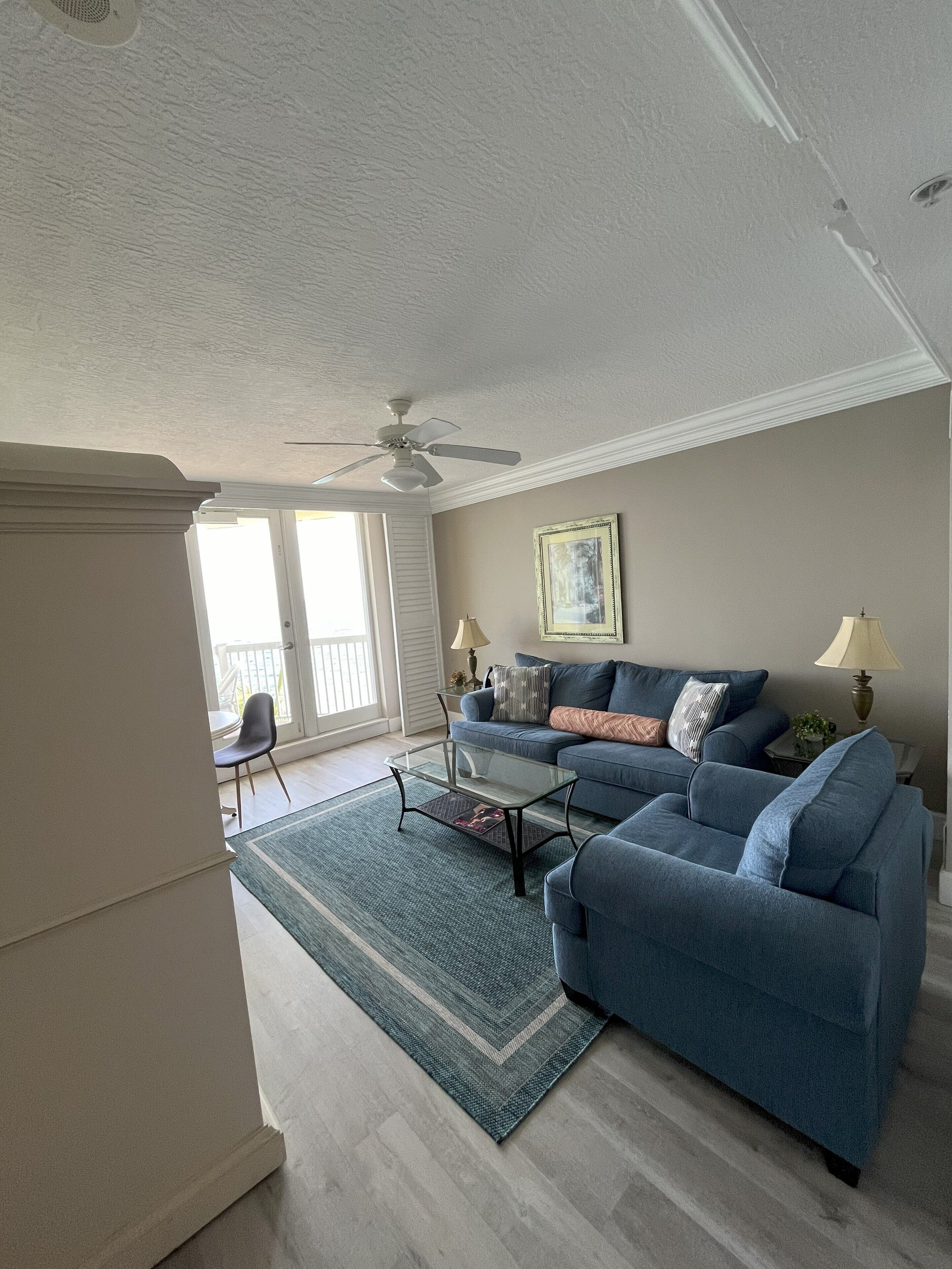 A living room with a blue sofa, a glass coffee table, a patterned rug, and a ceiling fan.
