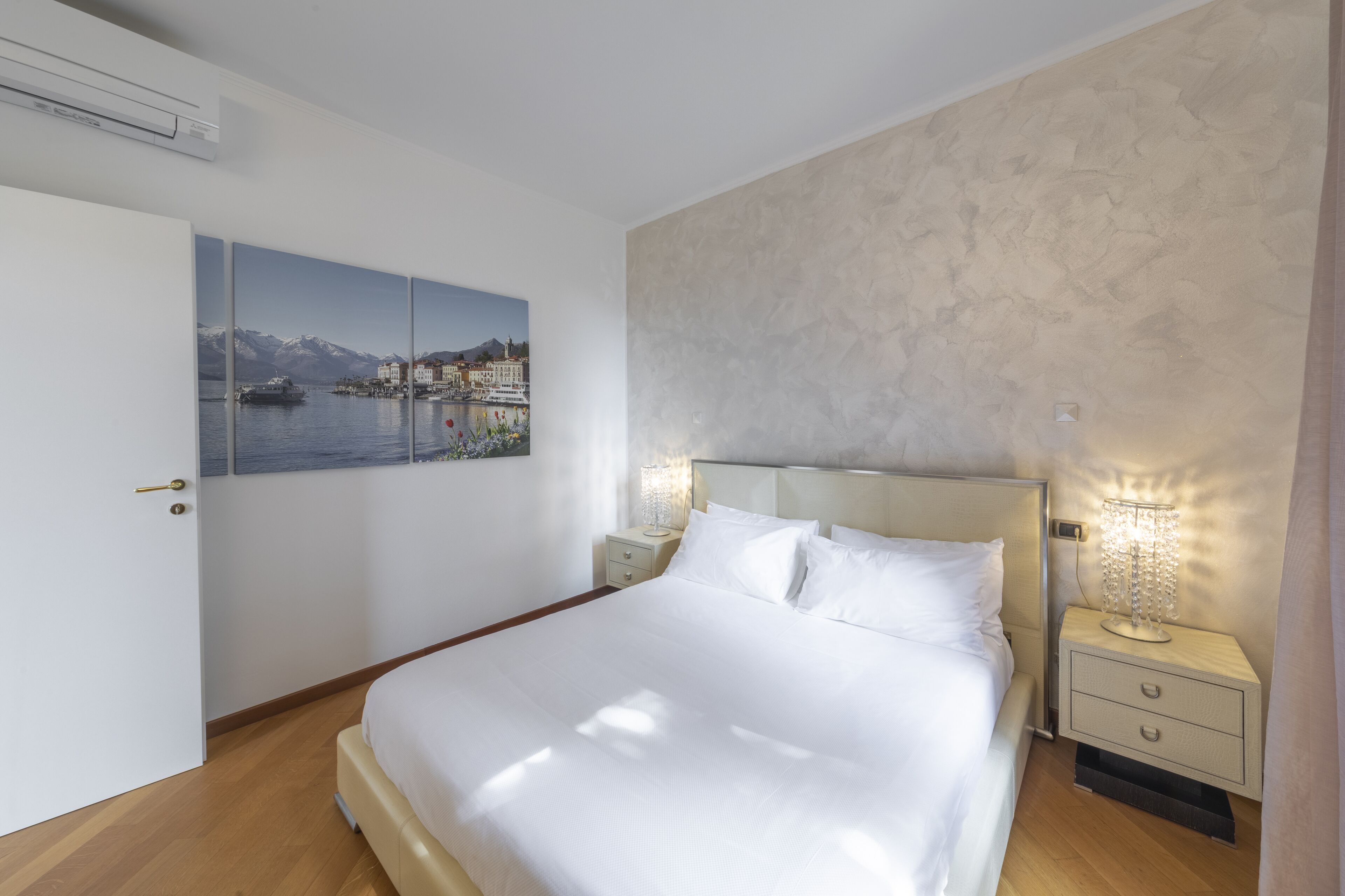 A bedroom with a bed, bedside tables, a nightstand, a wall-mounted air conditioner, and a framed picture of a lakeside scene.