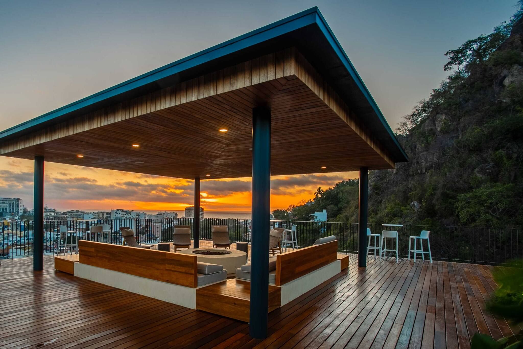 Un espace extérieur abrité, aménagé avec des sièges, offrant une vue sur les toits d’une ville au coucher du soleil.