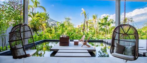A serene outdoor seating area with a pool, surrounded by lush greenery and a clear blue sky.