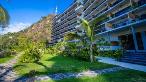 A multi-story hotel with balconies, a garden area, and a view of a hill with a tower in the background. - Casa Costera | Walk to Romantic Zone & Beach (Puerto Vallarta)