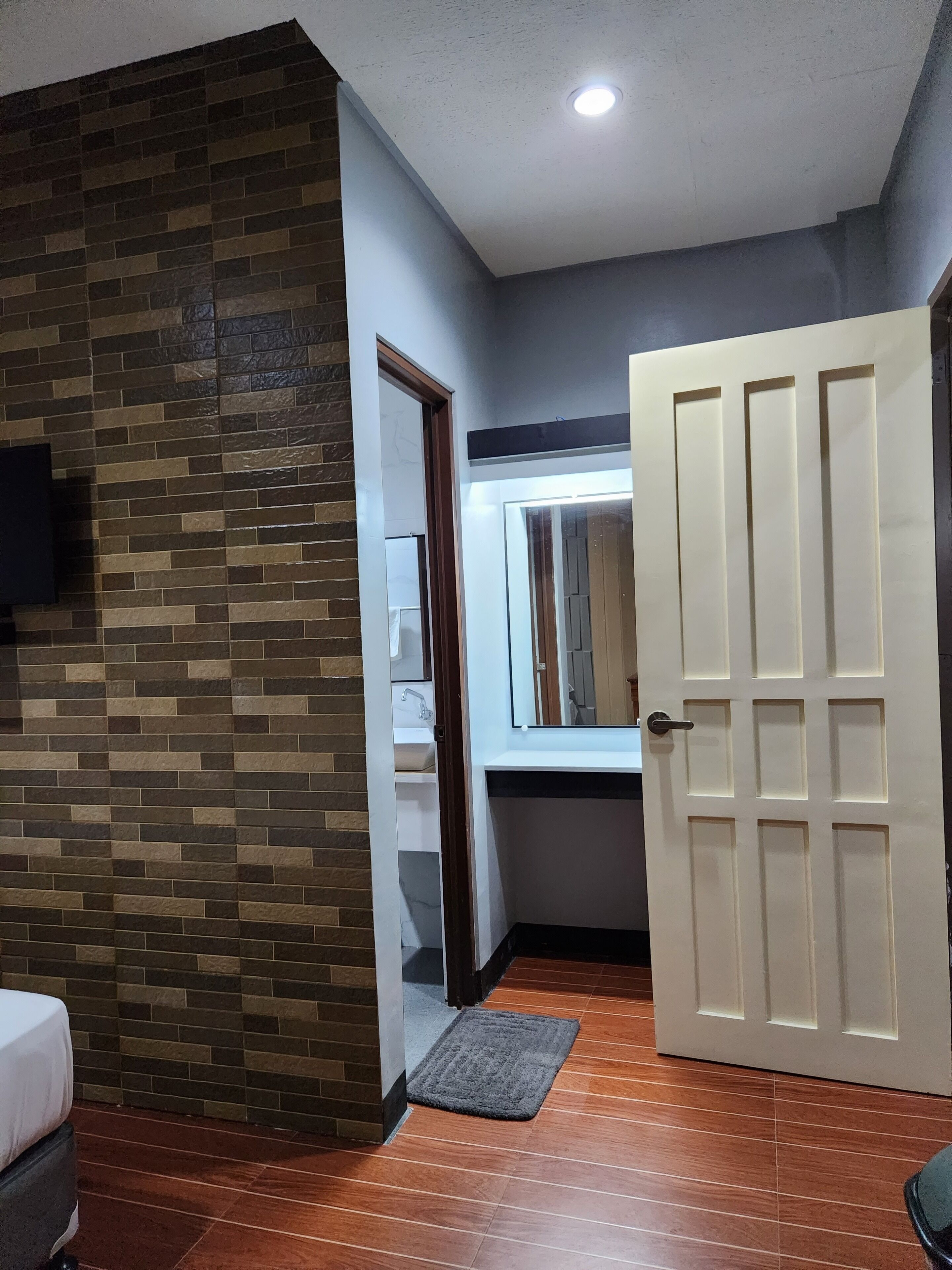 A room with a brick wall, a white door, a mirror, and a small bathroom with a sink and mirror.