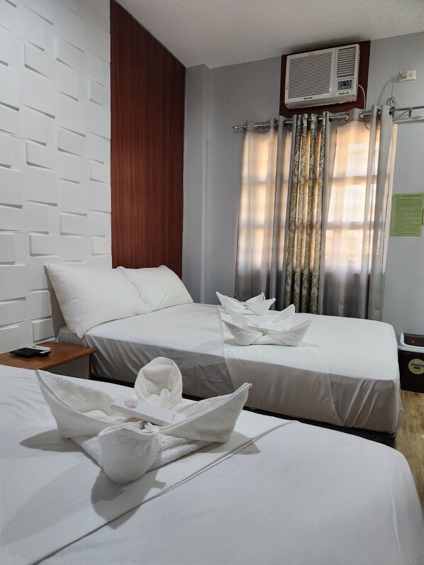 Une chambre d’hôtel avec deux lits, une tête de lit en bois, un appareil de climatisation, une table de chevet avec une télécommande et une poubelle.