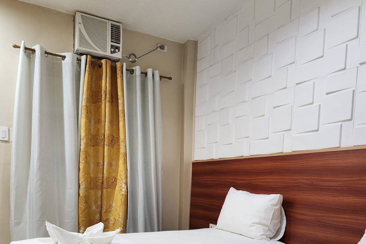 Ein Hotelzimmer mit zwei Betten, jeweils mit weißer Bettdecke und einem ordentlich gefalteten Handtuch. Auf einer Seite ist ein hölzernes Kopfteil, auf der anderen eine strukturierte weiße Wand zu sehen.
