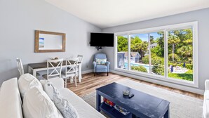 Een woonkamer met een groot raam, een flatscreen televisie, een eethoek en uitzicht op een zwembad en groen.