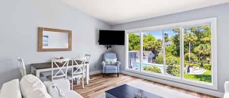 A living room with a large window, a flat-screen TV, a dining area, and a view of a pool and greenery.