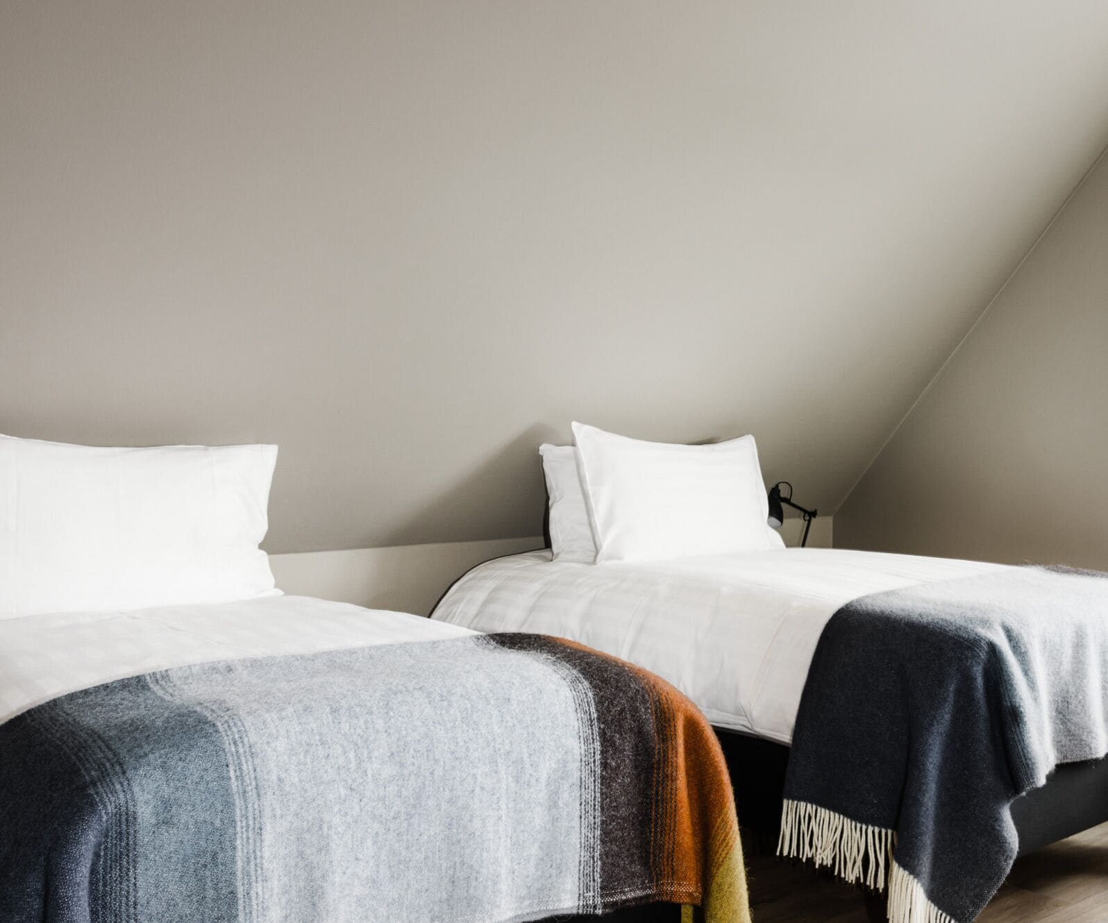 Dos camas con ropa blanca y un edredón oscuro sobre un piso de madera en una habitación con techo inclinado.