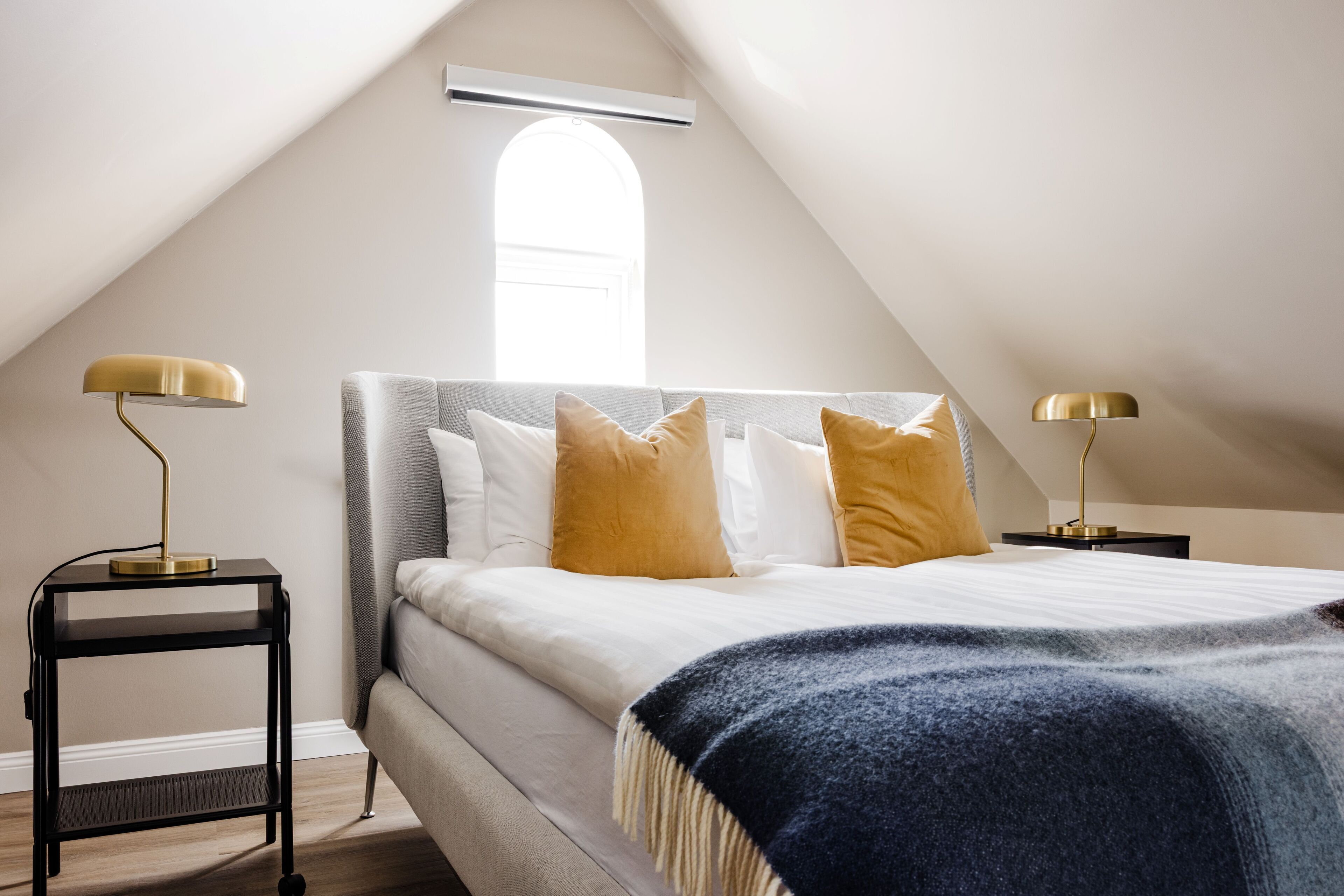 A bedroom with a sloped ceiling, a bed with white and yellow pillows, two bedside tables with lamps, and a window with natural light.