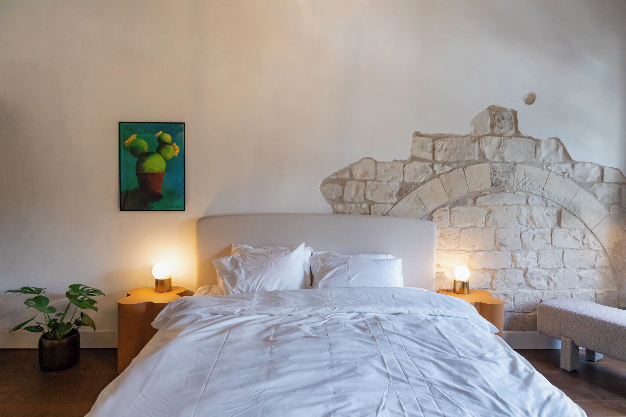 A bedroom with a stone wall, a bed with white bedding, two bedside tables with lamps, and a potted plant.