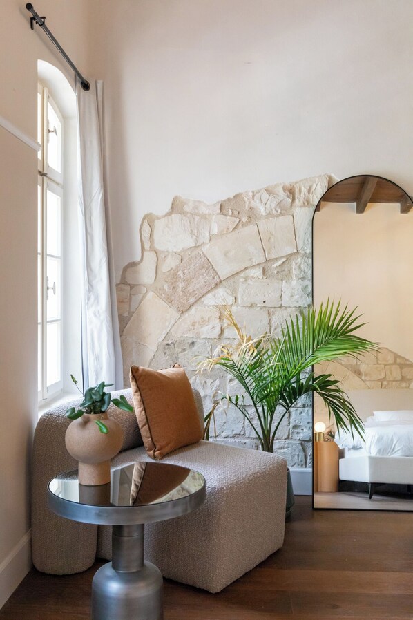 A cozy room with a stone wall, a round mirror, a sofa with cushions, a small round table, and a potted plant.