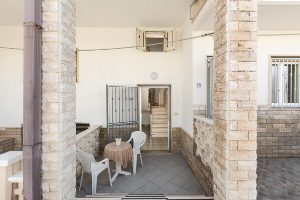 A small outdoor seating area with a table and two chairs, a stone pillar, and a door leading to an interior space.