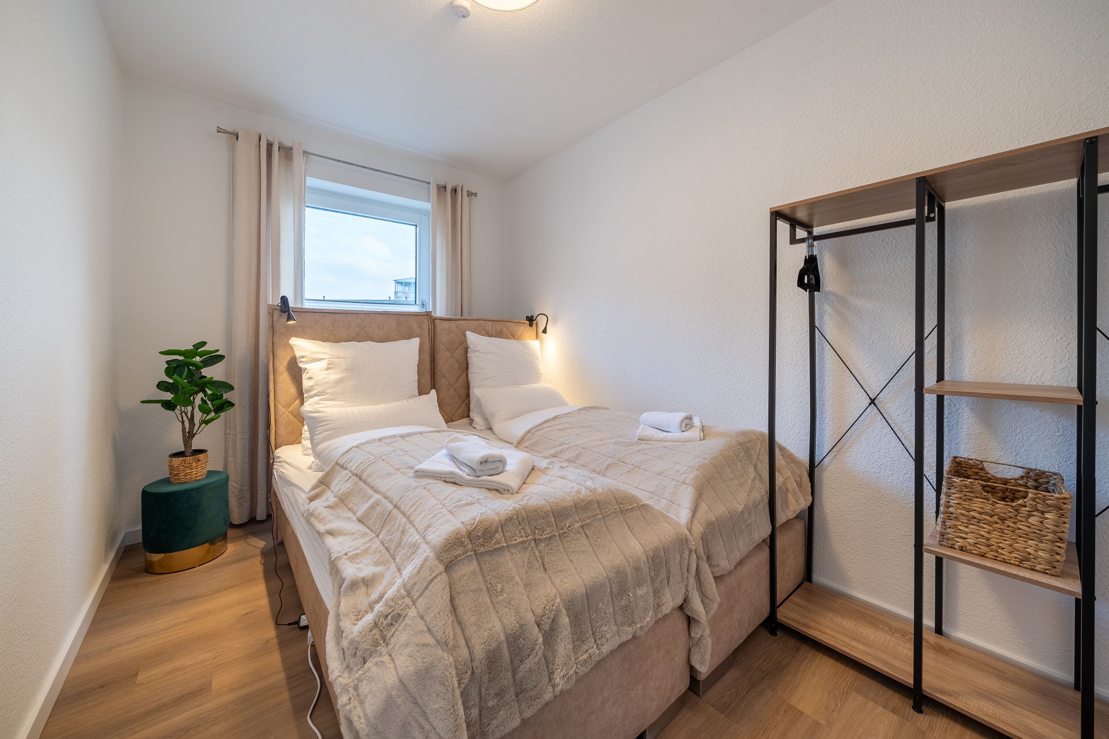 Ein Schlafzimmer mit einem Bett, einem Nachttisch mit Pflanze, einem Fenster mit Vorhängen und einem Regal mit Körben.