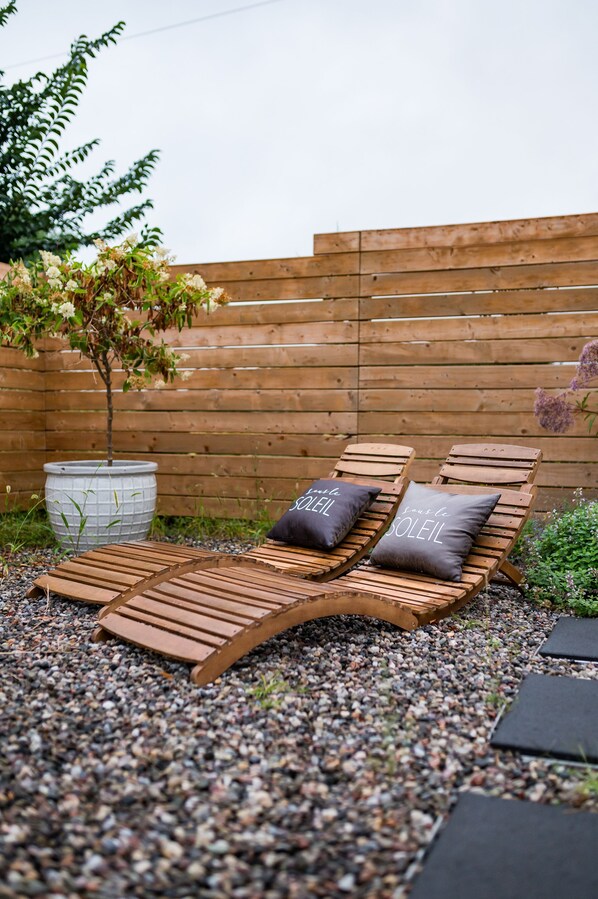 A wooden lounge chair with cushions, a large planter, and a wooden fence in a garden setting. - Lakecroft (Prince Edward)