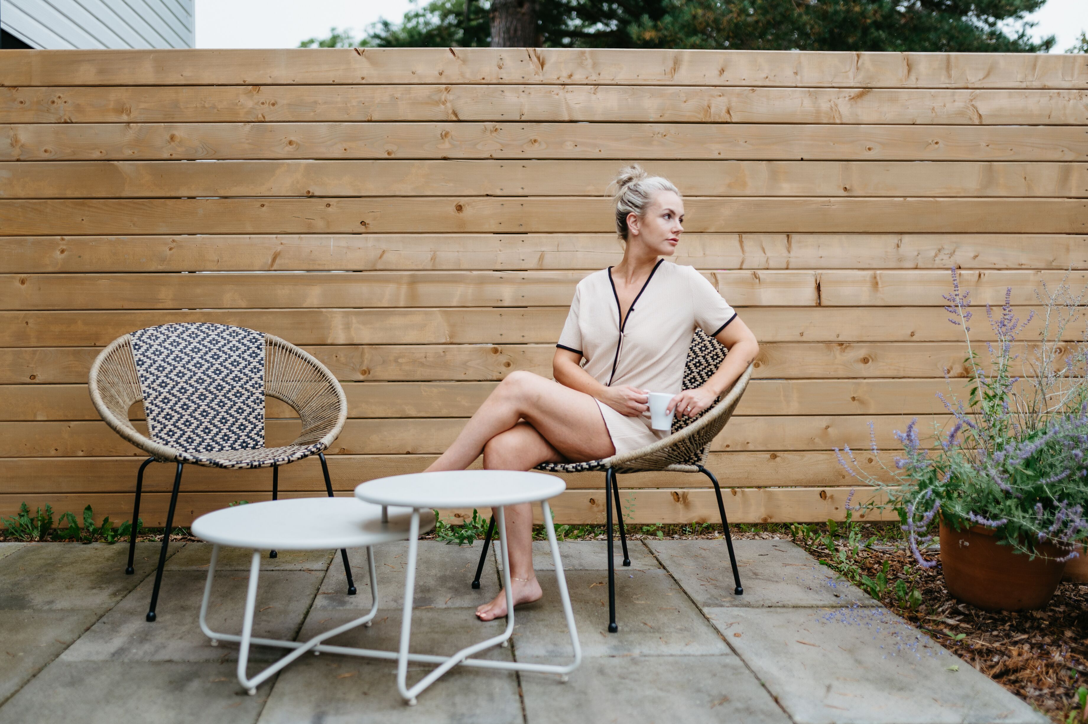 A person sitting on a wicker chair with a patterned cushion, holding a cup, next to a small round table.