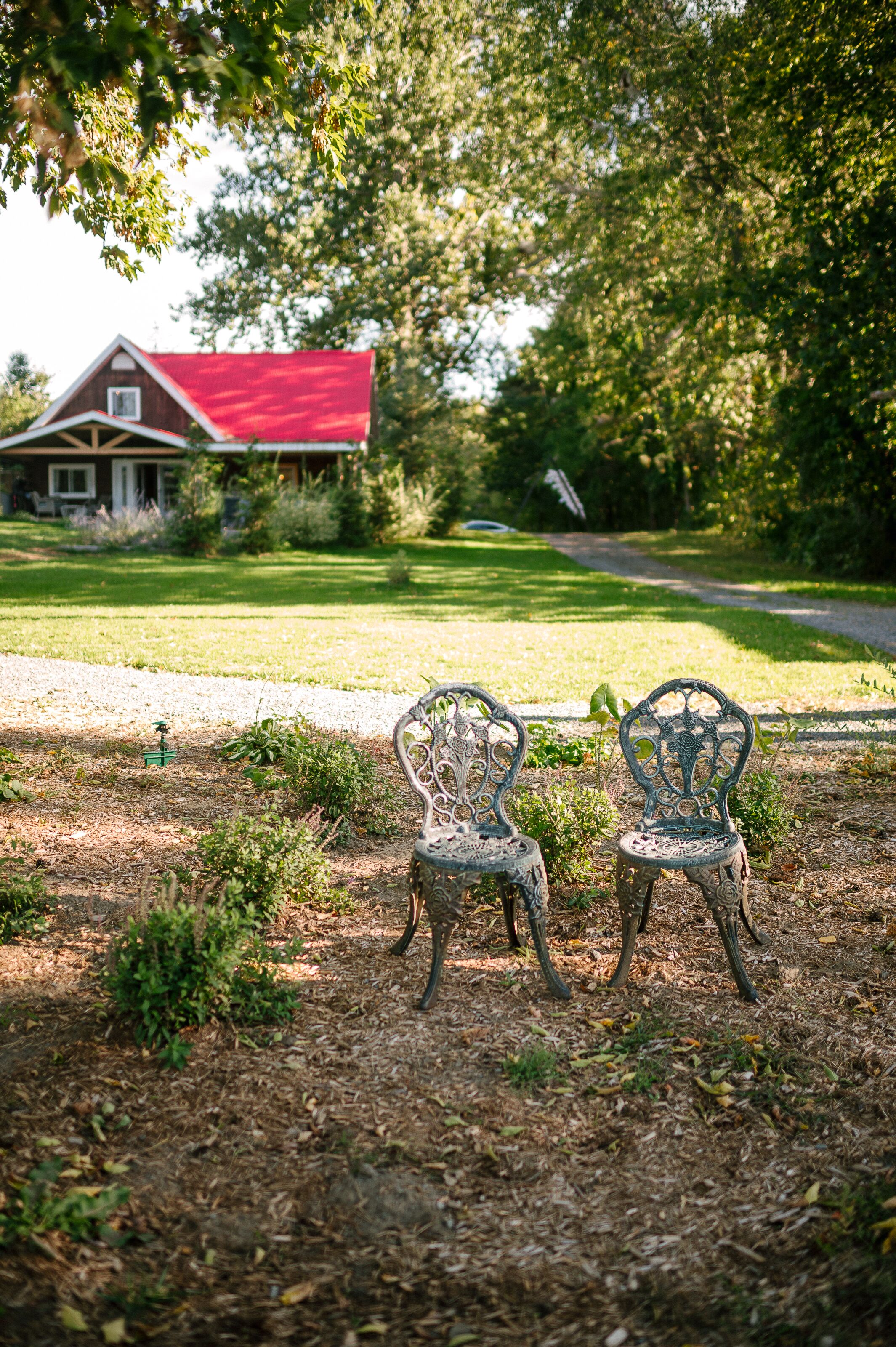 Deux chaises en métal richement décorées se trouvent dans un jardin, avec un bâtiment à toit rouge en arrière-plan.