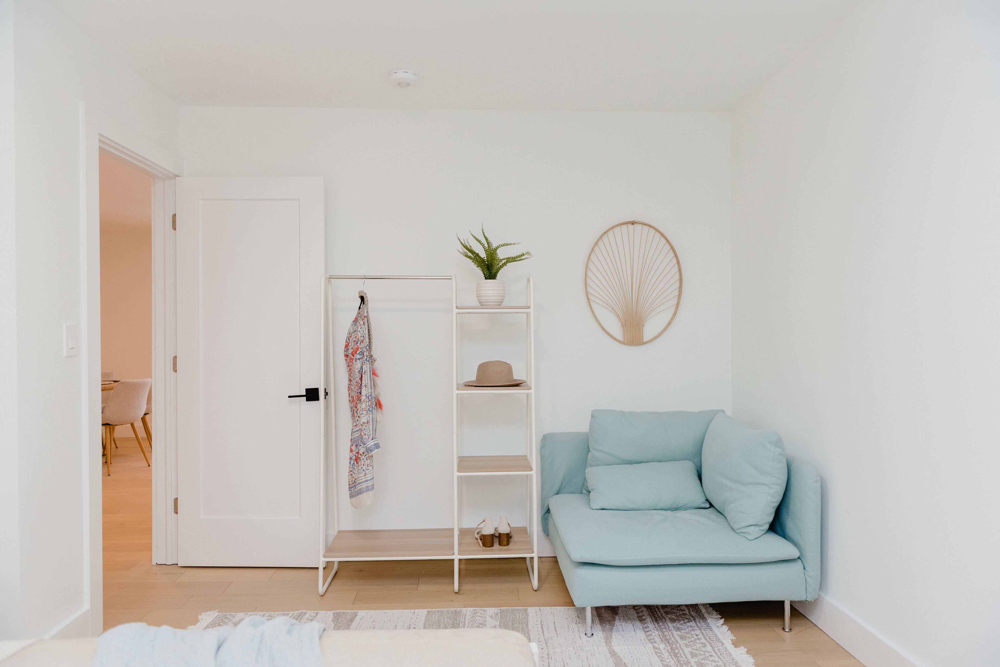 A minimalist living room with a light blue sofa, a white door, a white shelving unit with a plant, and a wall-mounted fan.