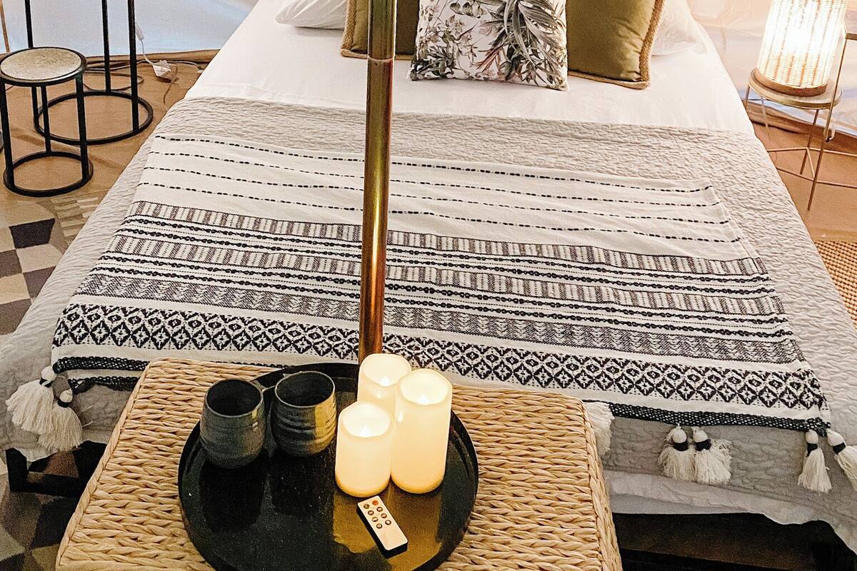 Habitación tipo alojamiento en tienda de campaña, con una cama, almohadas, una mesita de noche con lámpara, una mesa tejida con velas y una bandeja con saleros y pimenteros.