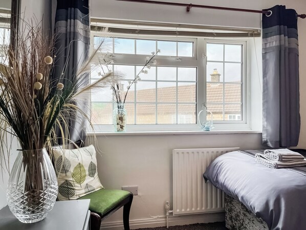 Un dormitorio con cama, radiador, ventana con cortinas, un florero con plantas secas y una silla.