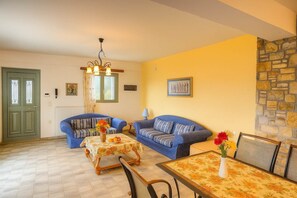 A living room with two blue sofas, a wooden coffee table, a dining table with a floral tablecloth, a chandelier, and a stone wall.
