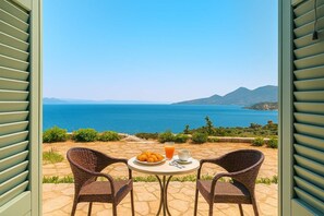 Eine offene Terrasse mit Korbstühlen, einem Tisch mit Obst und einem Glas Orangensaft, mit Blick auf das Meer und die Berge.