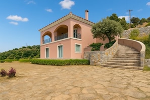 A pink villa with a stone staircase leading to a balcony, surrounded by greenery and a stone wall.