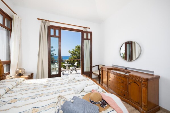 Un dormitorio con cama, cómoda, espejo redondo y balcón con vista al mar.
