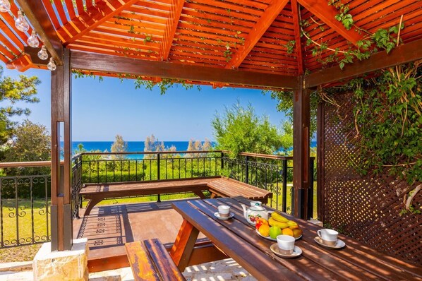 An outdoor seating area with a wooden table, chairs, and a view of the sea. - Villa Ariadne - Four Bedroom Villa, Sleeps 10 (Argaka)
